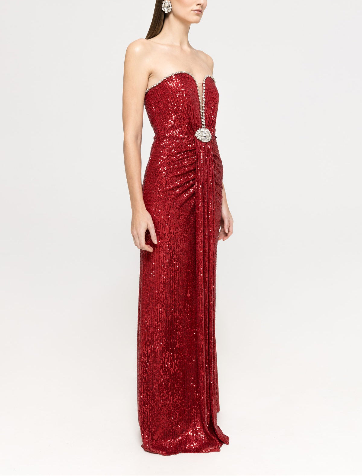 Side view of model wearing a strapless red sequined gown with a deep V crystal‑trim neckline, ruched waist and a draped detail, revealing the figure‑hugging silhouette.