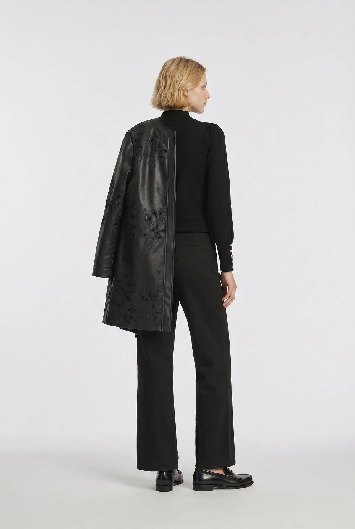 Weill Paris black floral embroidered faux leather coat draped over the model's shoulder with a black trouser outfit.