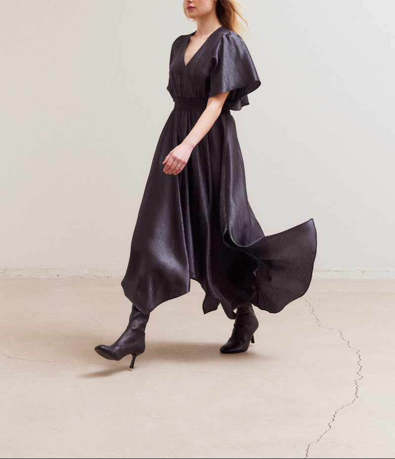 Model walking in flowy handkerchief-hem satin dress and heeled boots in a minimalist setting.