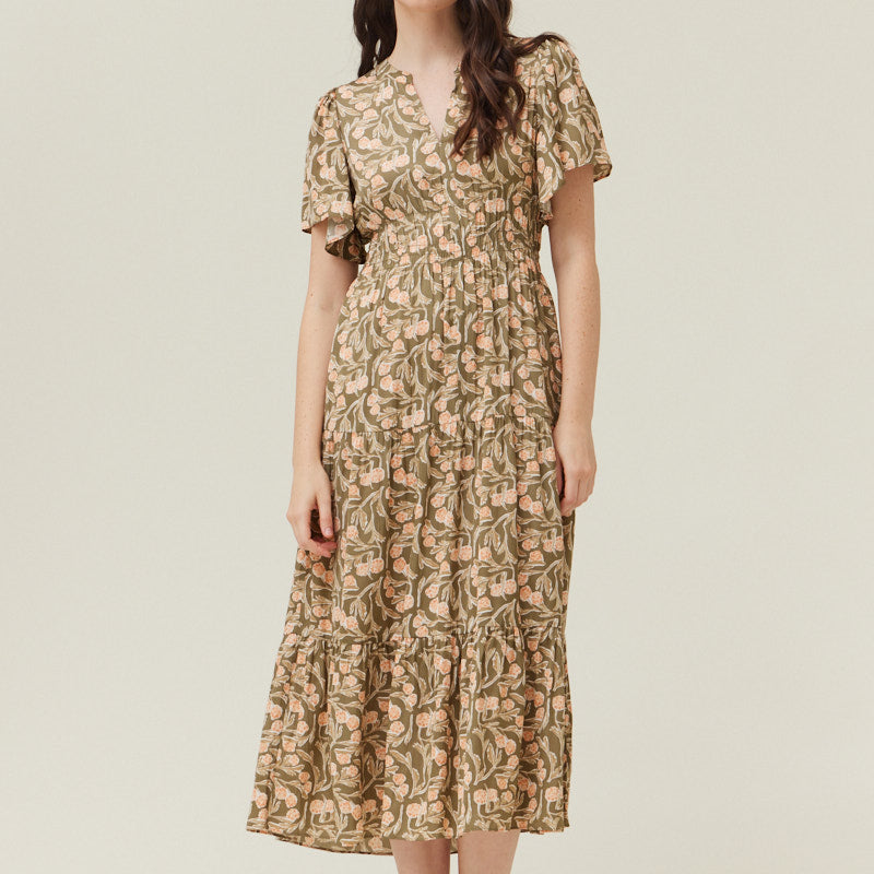 A woman standing in a studio setting, wearing a floral midi dress with short flutter sleeves, a V-neckline, and a tiered skirt.
