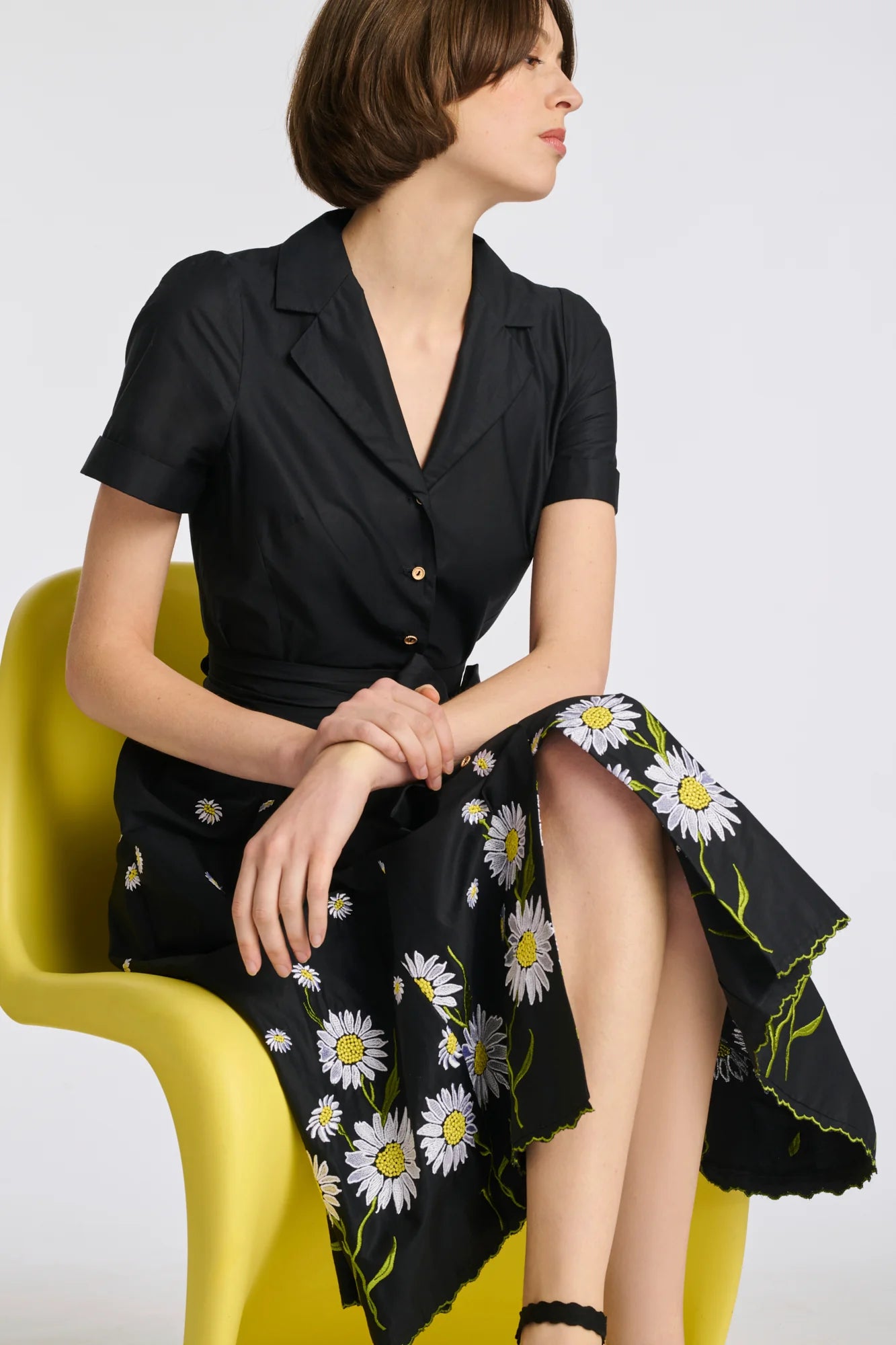 Model seated in black embroidered cotton midi shirt dress with floral hem embroidery and tie belt