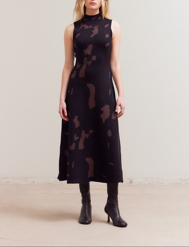 A model wearing a sleeveless black and brown abstract patterned knit maxi dress with a high mock neckline.