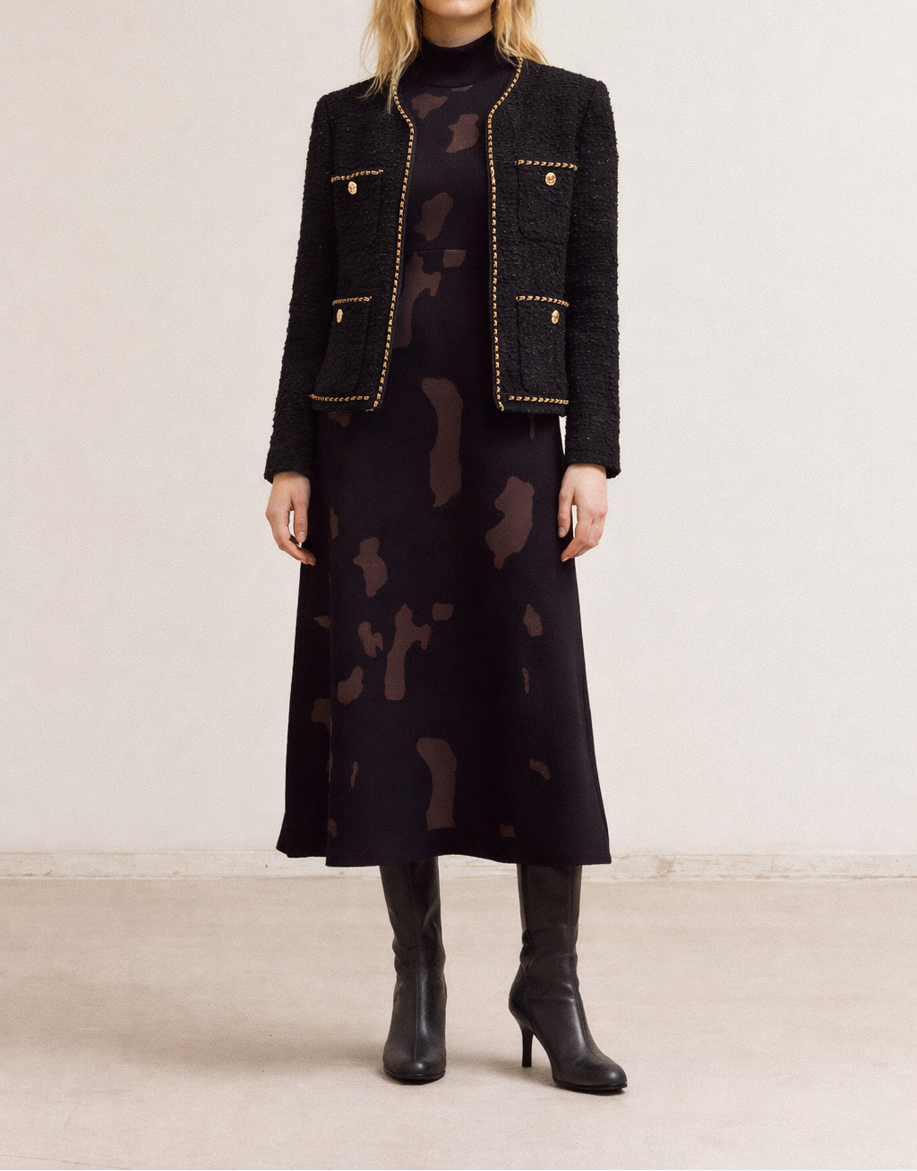 Woman wearing a black tweed jacket with gold chain trim and black fitted knit maxi dress on a plain background