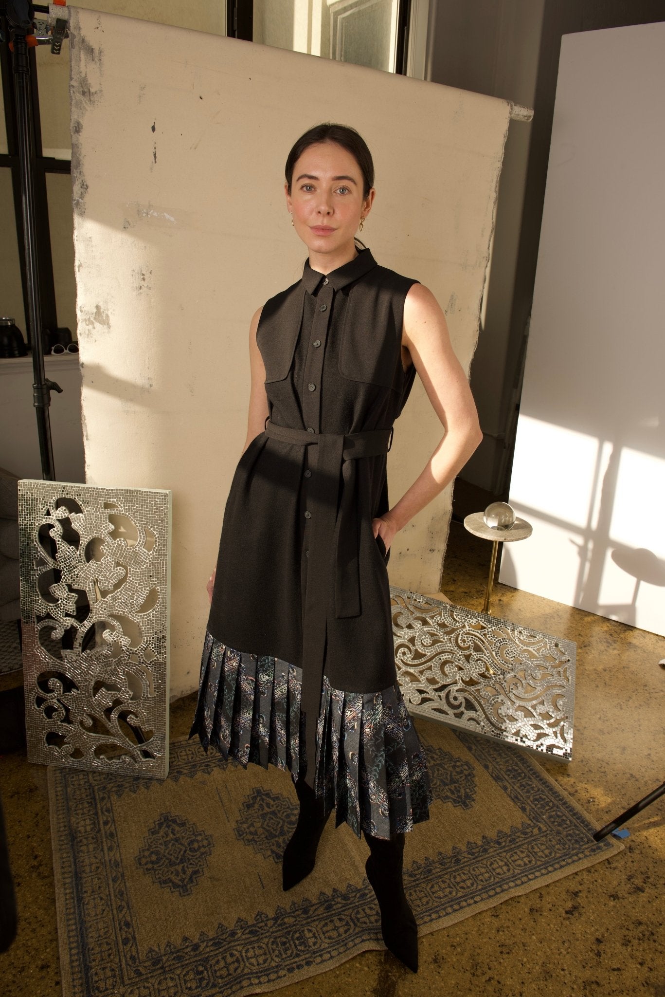 A woman standing in a studio setting, wearing a black Jelda Midi Dress with a collared neckline, button-down front, and a belted waist. The dress features a pleated floral hemline and is accessorized with a black belt and black ankle boots.