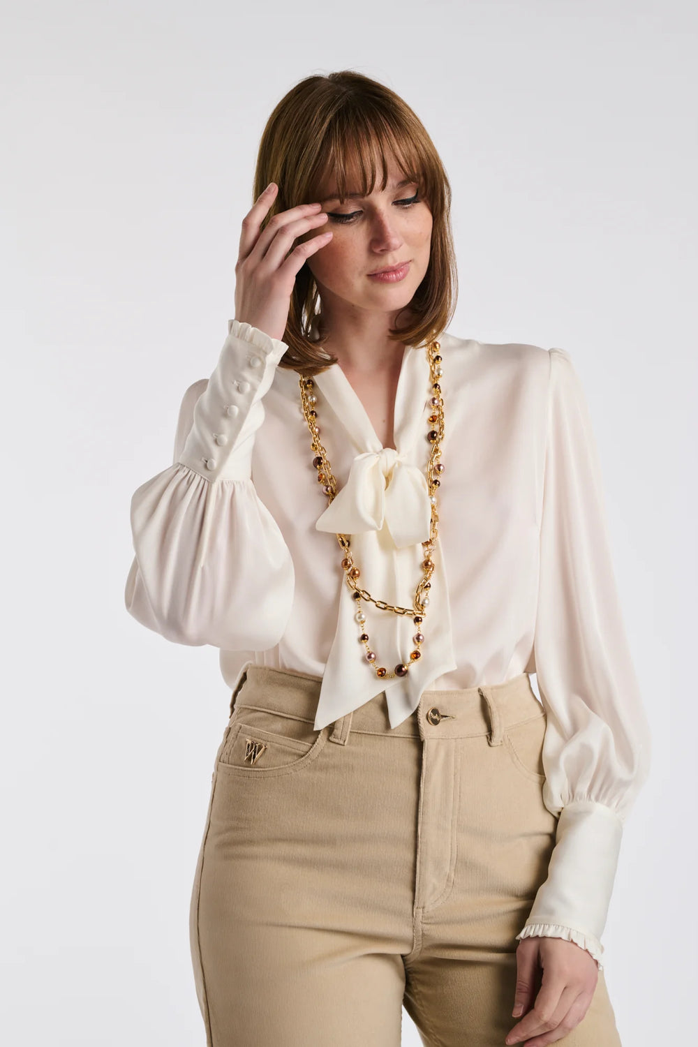 Woman wearing a long double‑chain gold‑tone necklace with brown and pearl‑effect beads over a cream bow‑neck blouse and beige high‑waisted trousers.