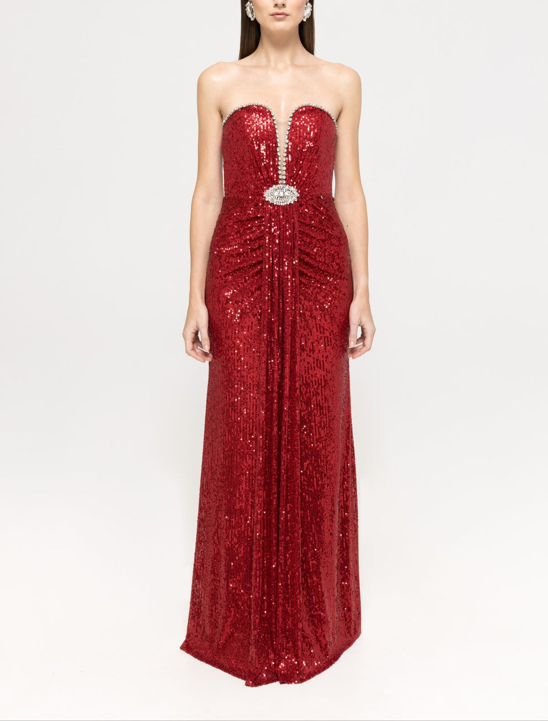 A model is wearing a strapless red sequined maxi dress with a crystal‑embellished plunge neckline and jeweled brooch at the waist, standing against a plain light background.		