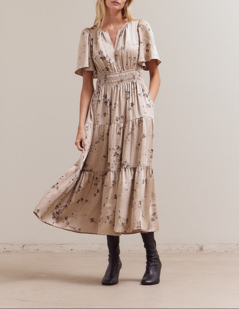 Front three‑quarter view of a natural‑beige floral tiered midi dress with V‑notch neckline, flutter sleeves, smocked waist and pockets, styled with black boots.