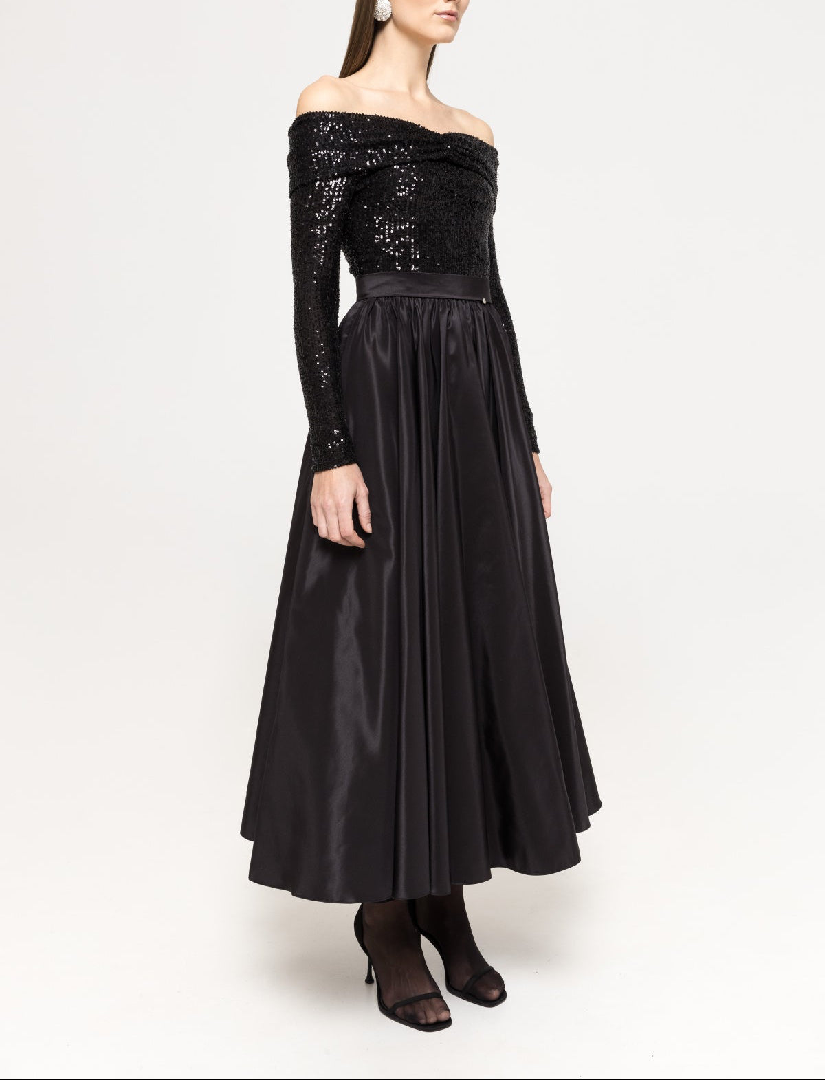 Three‑quarter angle of a black sequin‑bodice, off‑shoulder long‑sleeve dress with a voluminous taffeta A‑line midi skirt and defined waist.