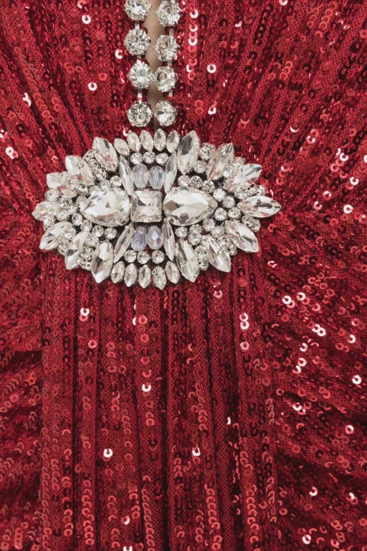 A model is wearing a strapless red sequined maxi dress with a crystal‑embellished plunge neckline and jeweled brooch at the waist