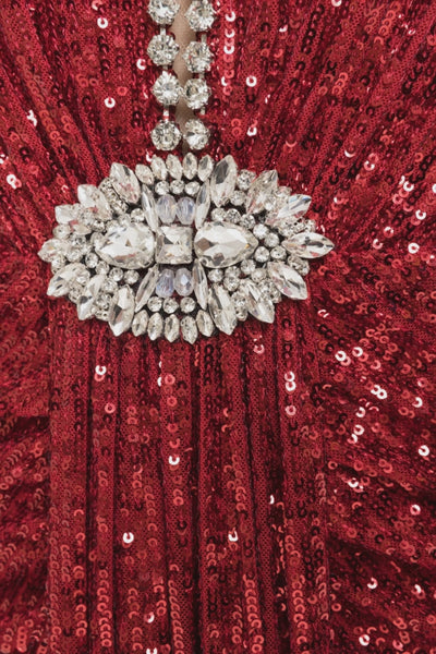 A model is wearing a strapless red sequined maxi dress with a crystal‑embellished plunge neckline and jeweled brooch at the waist