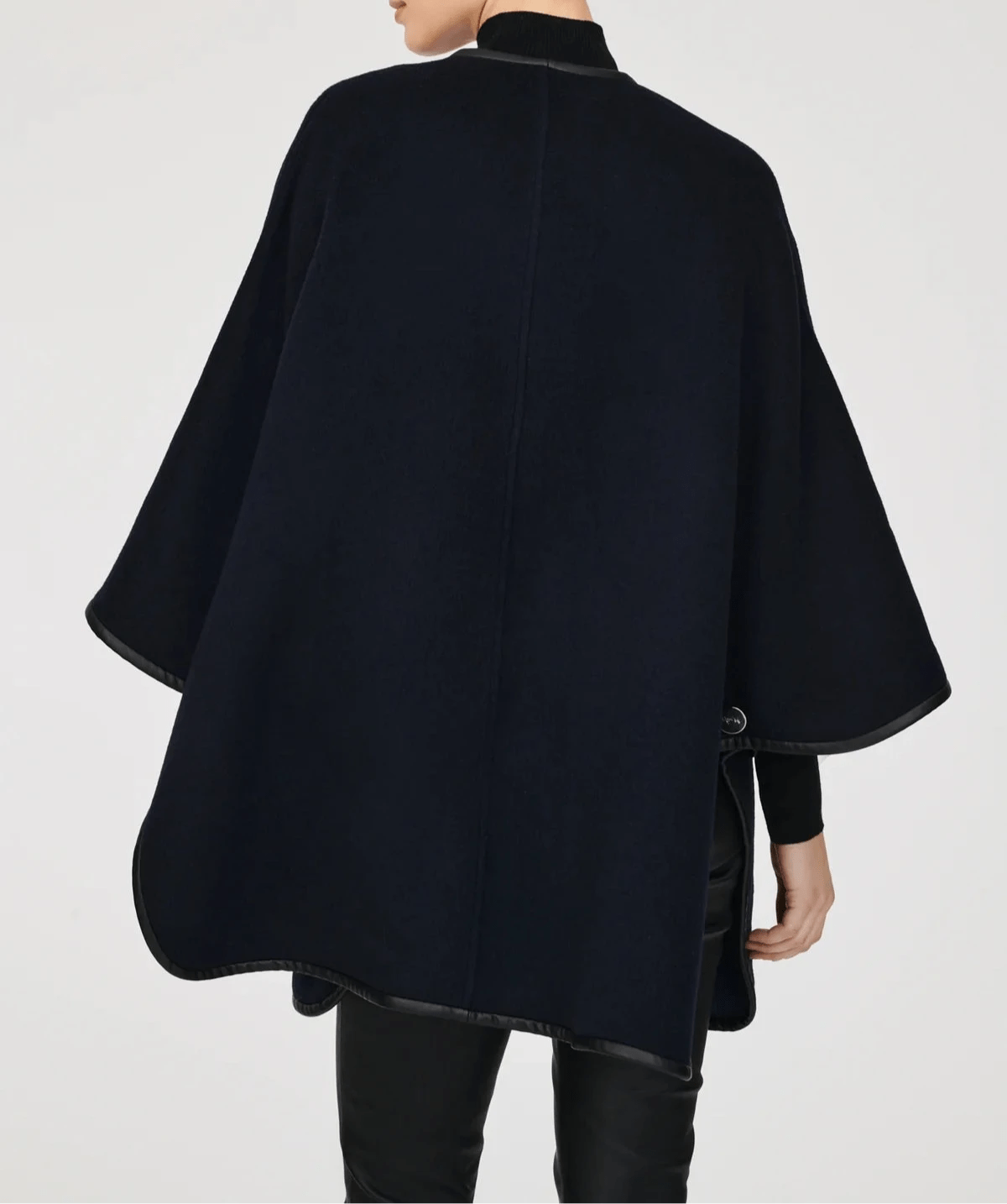 Back view of a model wearing the Weill Paris reversible wool cape in navy blue with black vegan leather trim, layered over a black turtleneck ensemble to create an elegant, minimalist winter look