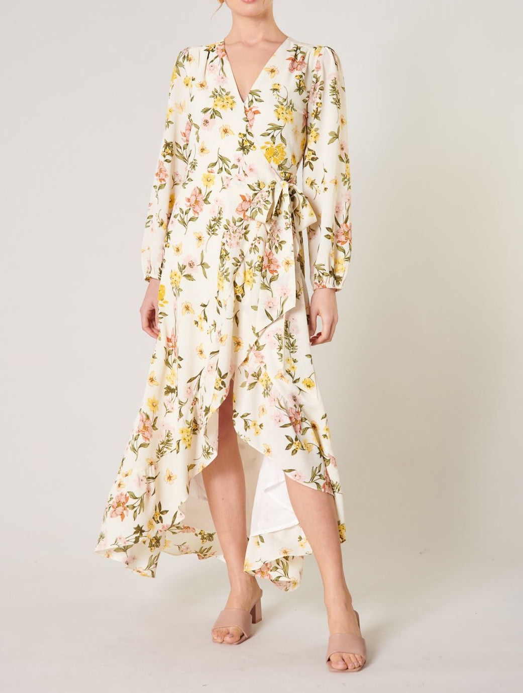 Model wearing the Ashlynn Floral Califa Maxi Wrap Dress by SUGARLIPS, featuring a flowing high-low hemline, long sleeves, and a vibrant print of yellow and pink flowers against a light cream background, complemented by beige strappy heels