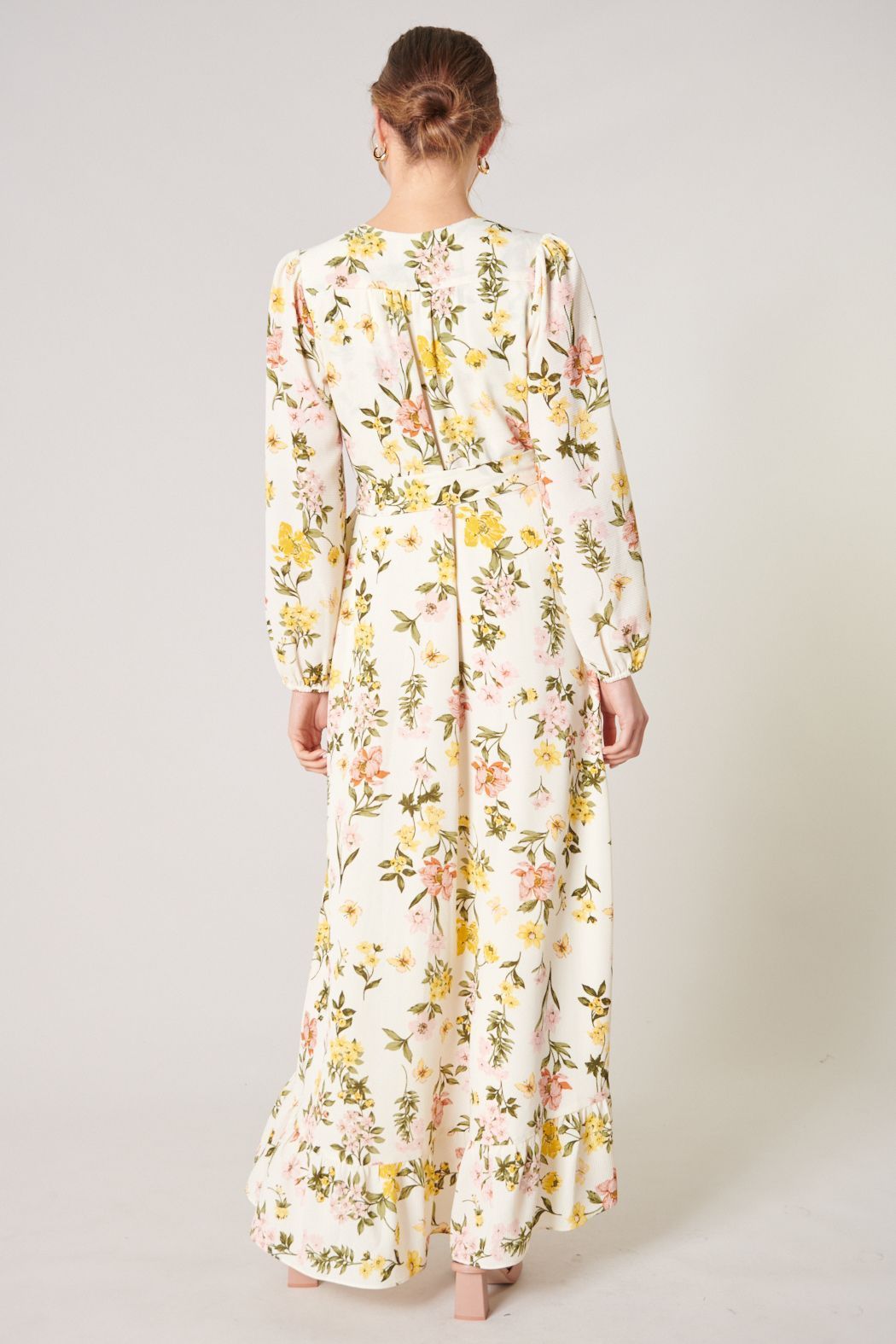 Model wearing the Ashlynn Floral Califa Maxi Wrap Dress by SUGARLIPS, featuring a flowing high-low hemline, long sleeves, and a vibrant print of yellow and pink flowers against a light cream background, complemented by beige strappy heels