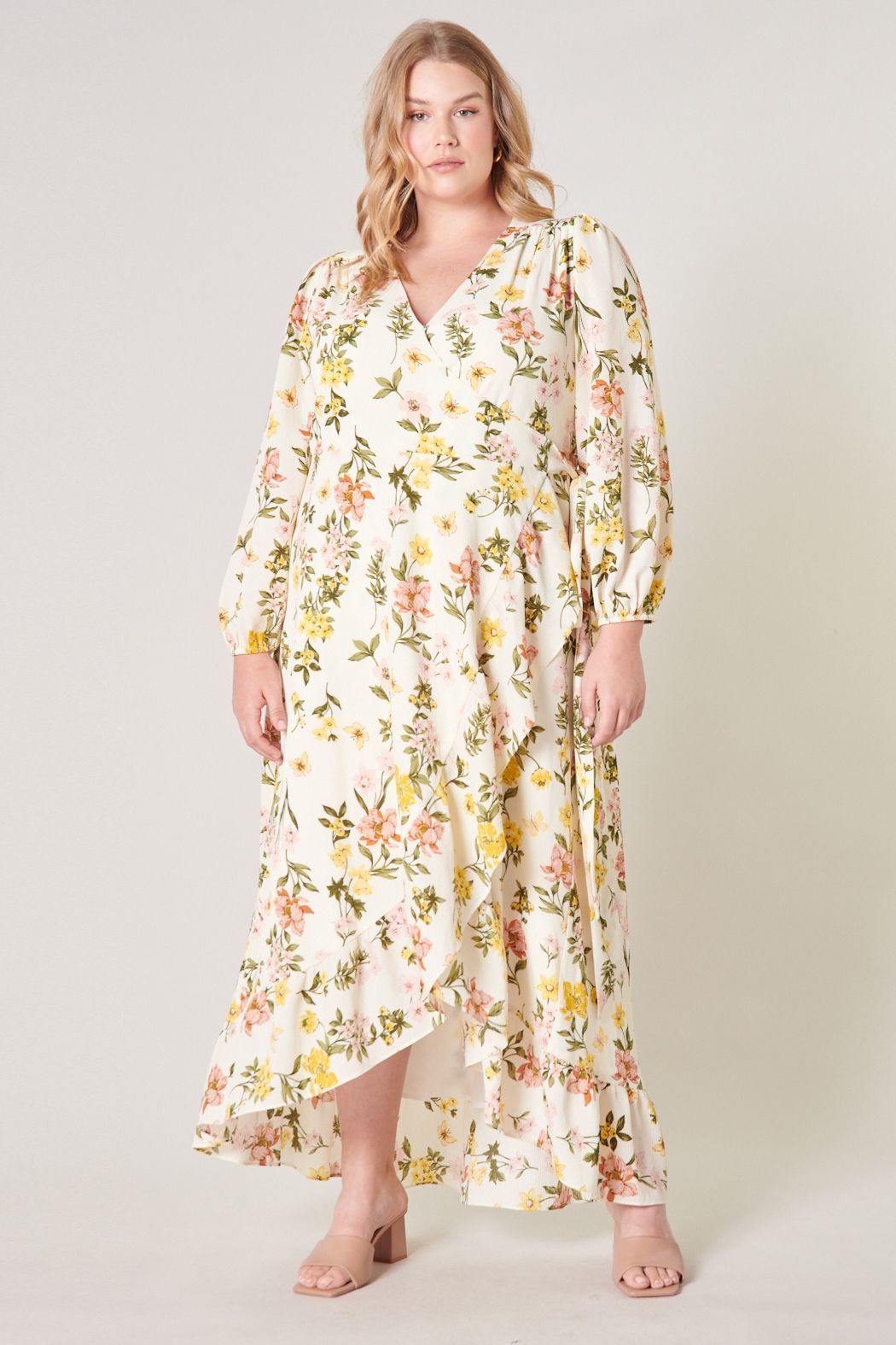 Plus Size Model wearing the Ashlynn Floral Califa Maxi Wrap Dress by SUGARLIPS, featuring a flowing high-low hemline, long sleeves, and a vibrant print of yellow and pink flowers against a light cream background, complemented by beige strappy heels