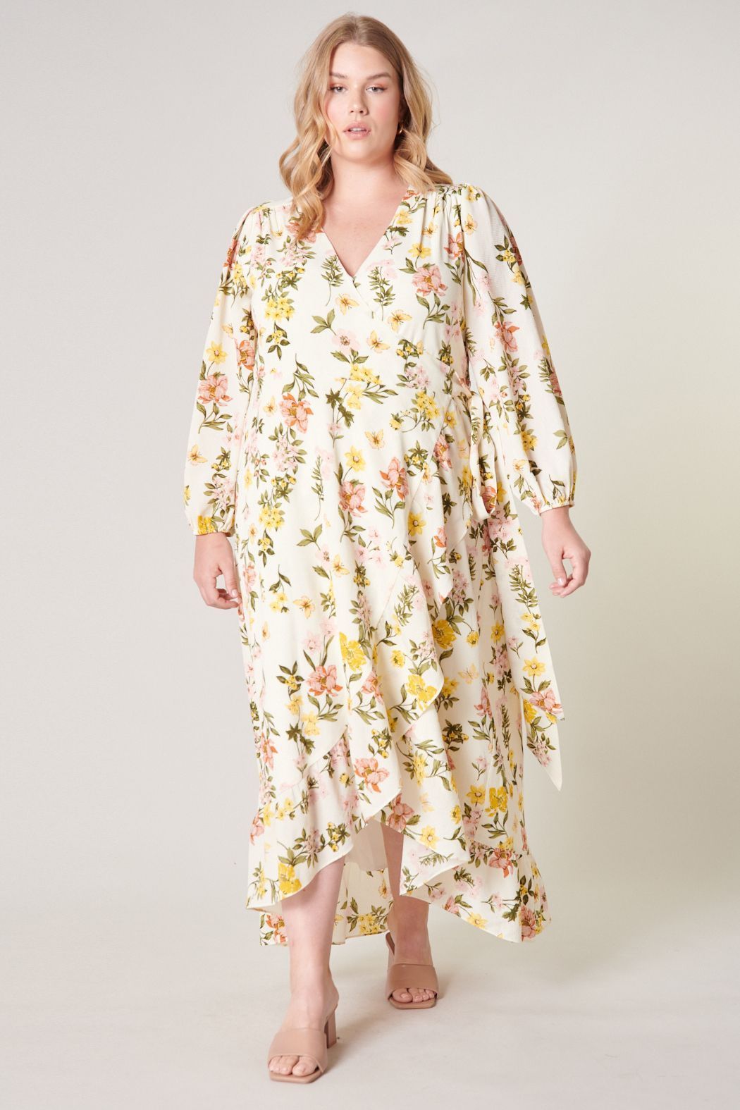 Plus Size Model wearing the Ashlynn Floral Califa Maxi Wrap Dress by SUGARLIPS, featuring a flowing high-low hemline, long sleeves, and a vibrant print of yellow and pink flowers against a light cream background, complemented by beige strappy heels